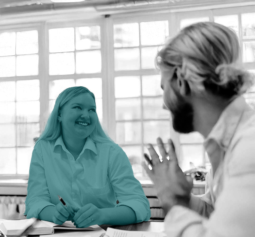 Eine Frau und ein mann sitzen am Besprechungstisch mit Schreibachen und lächeln sich an.