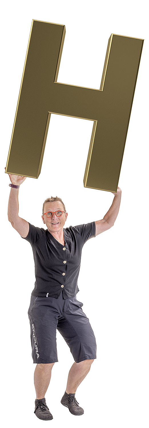 Barbara hebt die Arme und hält den Buchstaben H nach oben und lacht in die Kamera